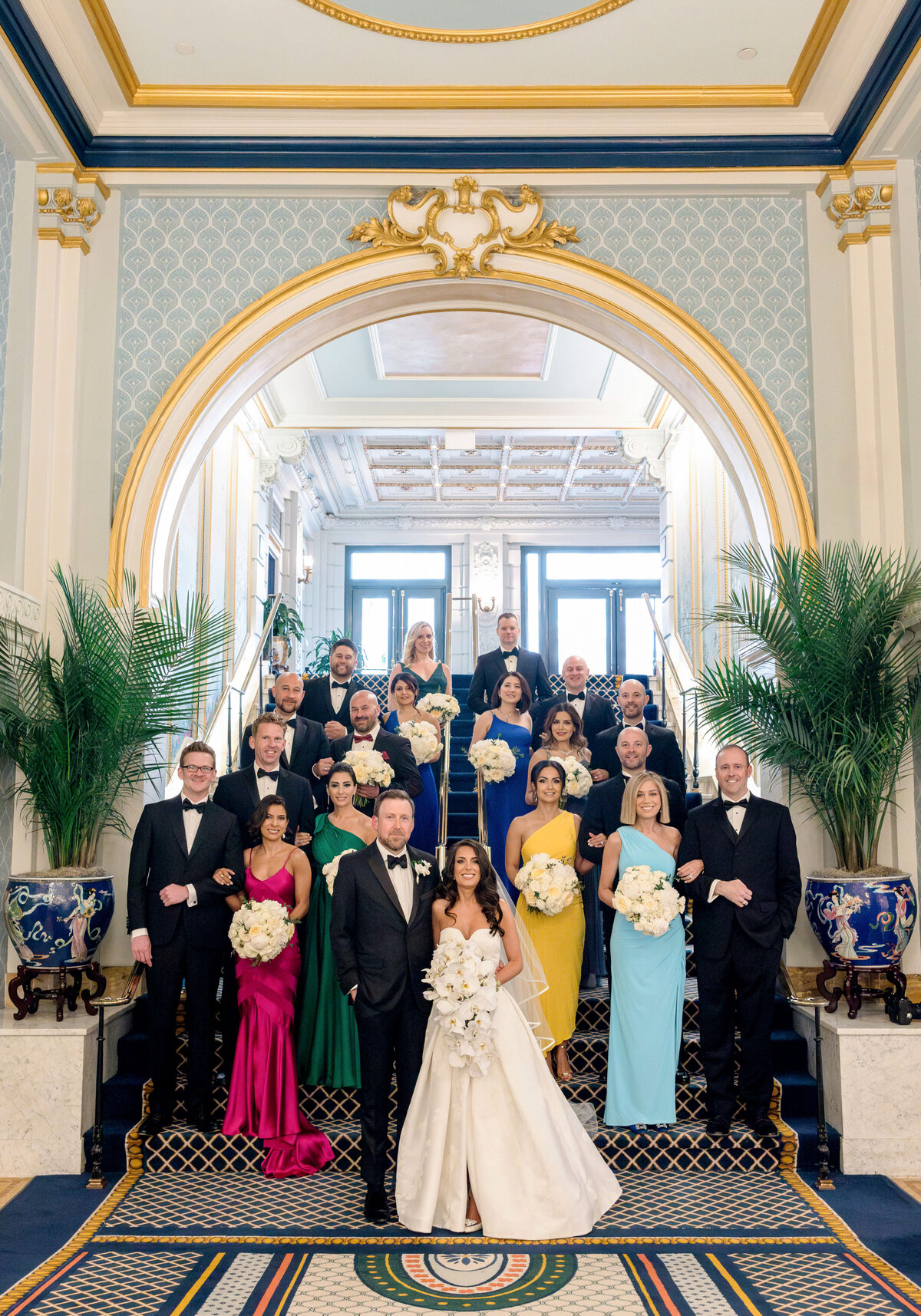 A large wedding party stands on an ornate staircase in formal attire. The bride and groom are at the front, surrounded by bridesmaids in colorful dresses and groomsmen in tuxedos, all holding bouquets. The decor features elegant columns and lush plants.