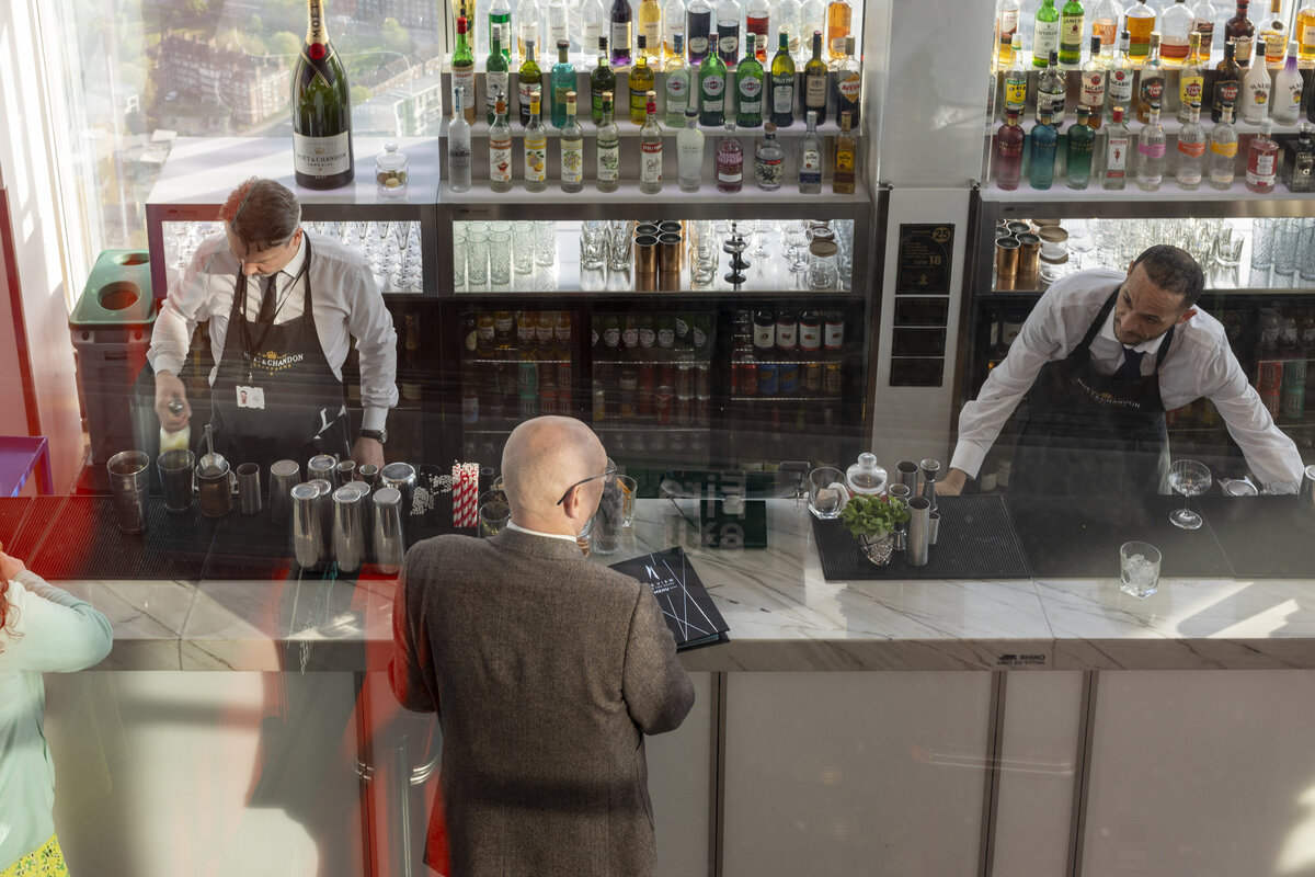 A couple of bartenders pictured taking drink orders from two customers. Photo is taken from a slightly elevated angle looking down own the scene unfolding. 
