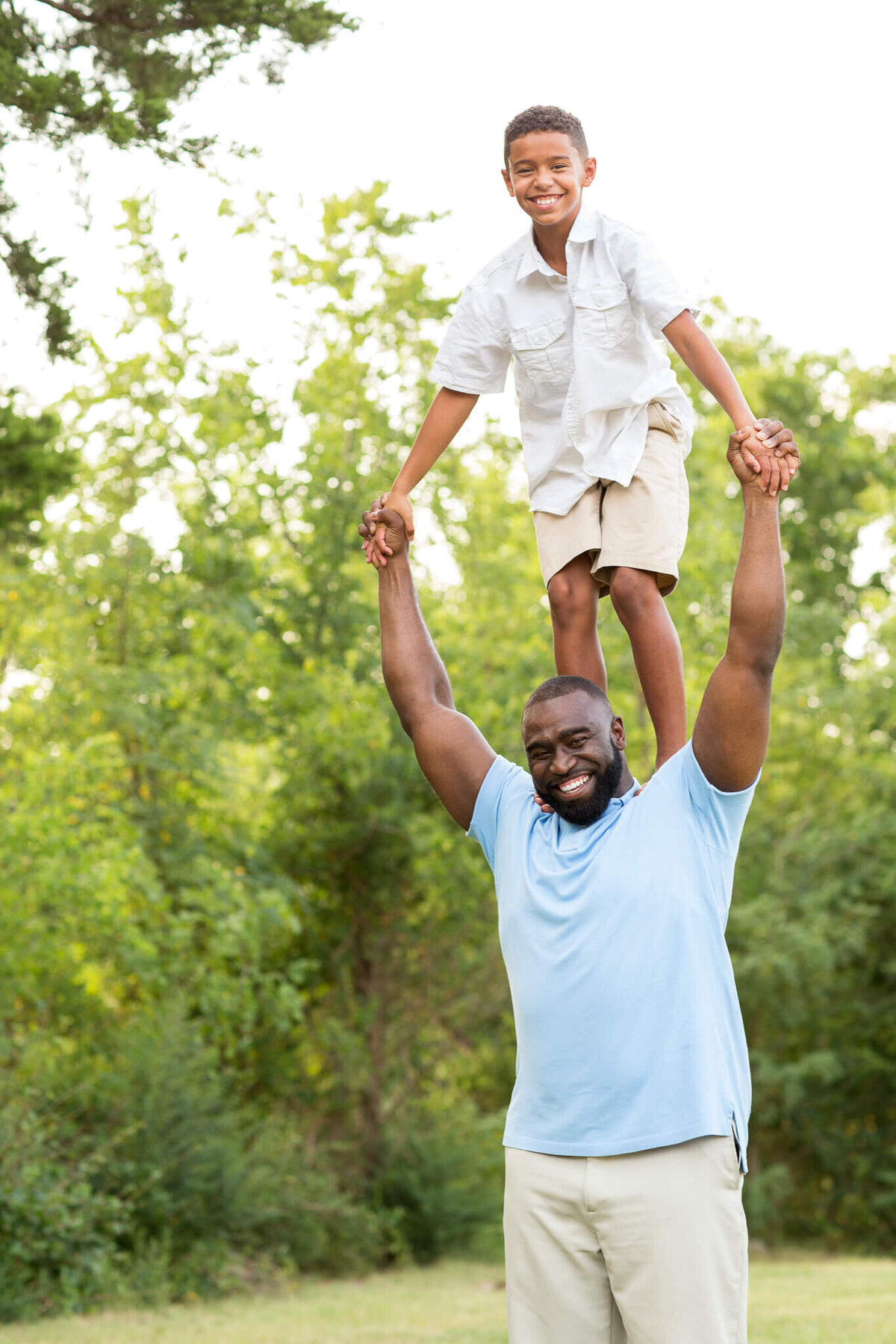 In a serene grassy area with trees, captured perfectly by a Fayetteville NC family photographer, a man in a light blue shirt joyfully lifts a smiling child onto his shoulders. They're holding hands as the child beams in their white shirt and beige shorts.