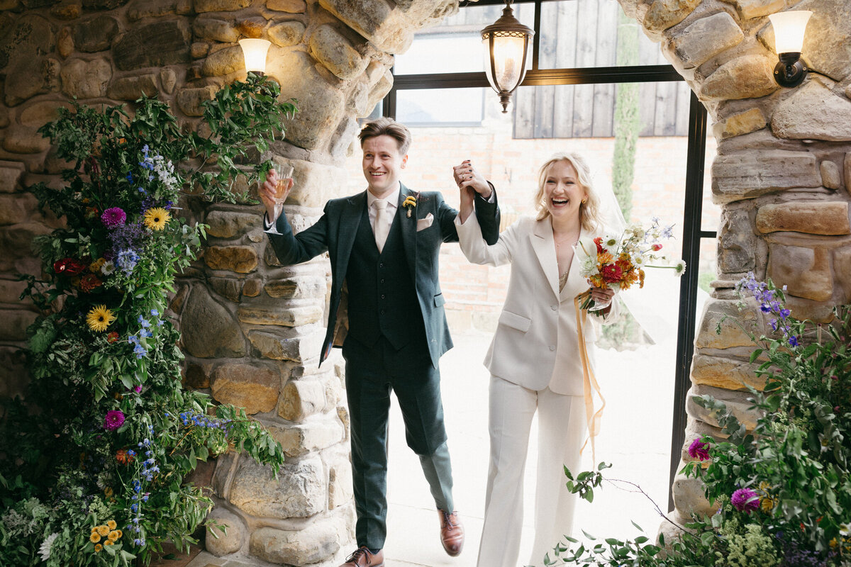 The bride and groom celebrate their newlywed status as they walk through a stone archway at Willow Marsh Farm. Both are smiling widely, holding hands with the groom raising a glass in one hand. The bride, in a chic white suit, holds a vibrant bouquet of flowers, while the groom is dressed in a green suit. The scene is framed by colorful floral arrangements on either side, creating a joyful and celebratory atmosphere.
