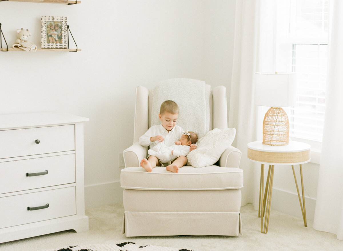 Big brother sister in a chair and holds baby sister during a Raleigh newborn session. Photographed by newborn photographer Raleigh A.J. Dunlap Photography.