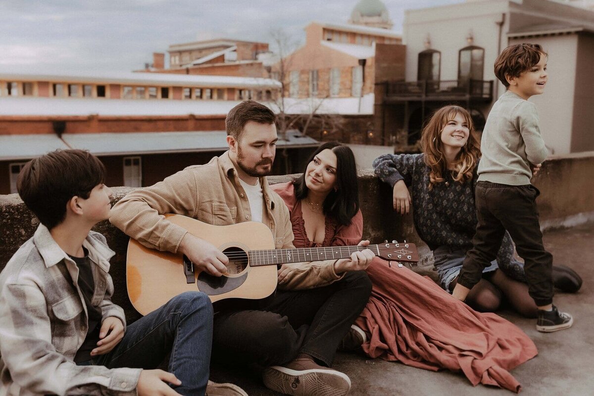 A group of people sits on a rooftop. One person plays a guitar, while others listen and relax. A young boy stands nearby. Urban buildings are in the background.