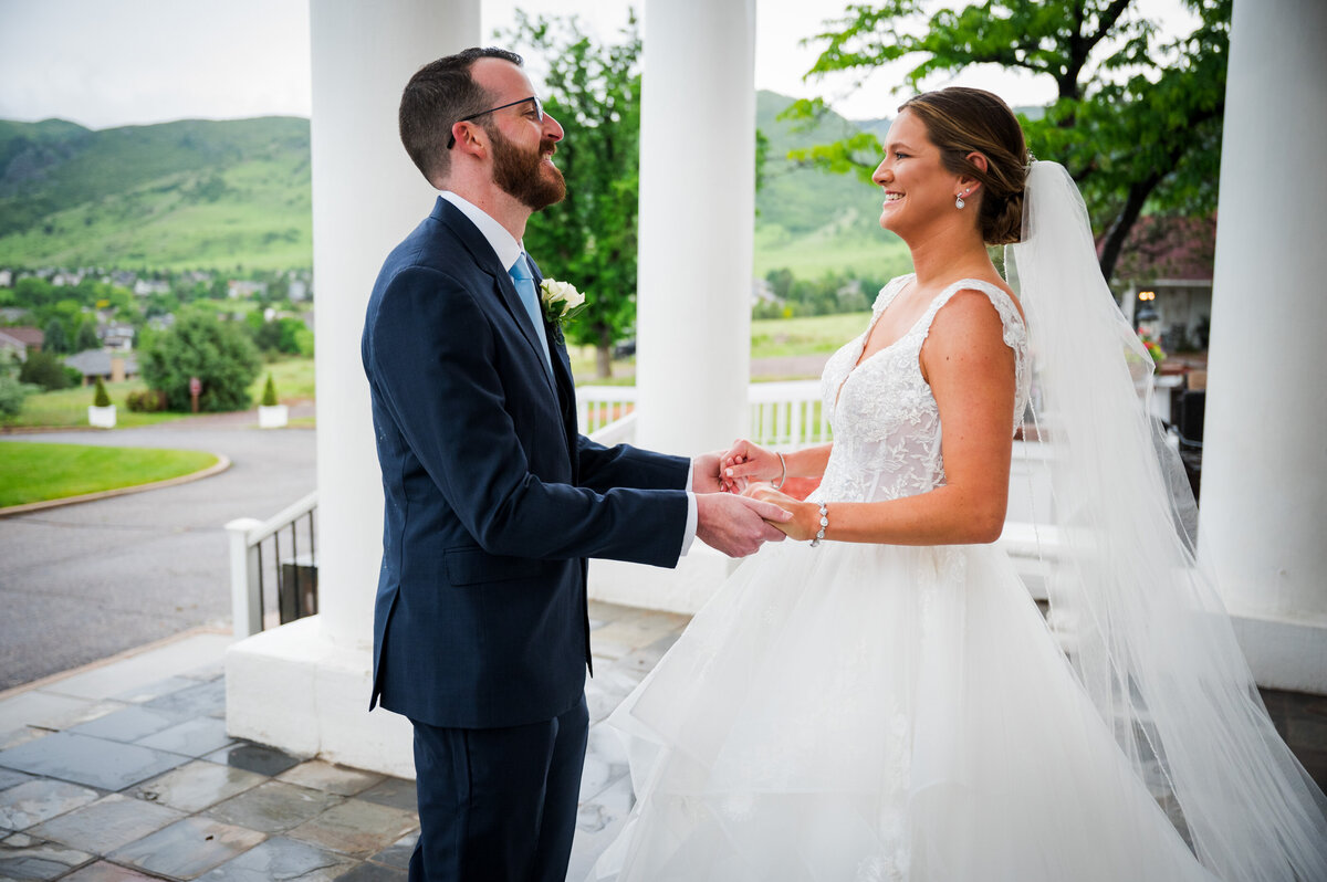 A bride and groom smile at each other and hold hands in reaction to their first look at The Manor House in Littleton, Colorado.