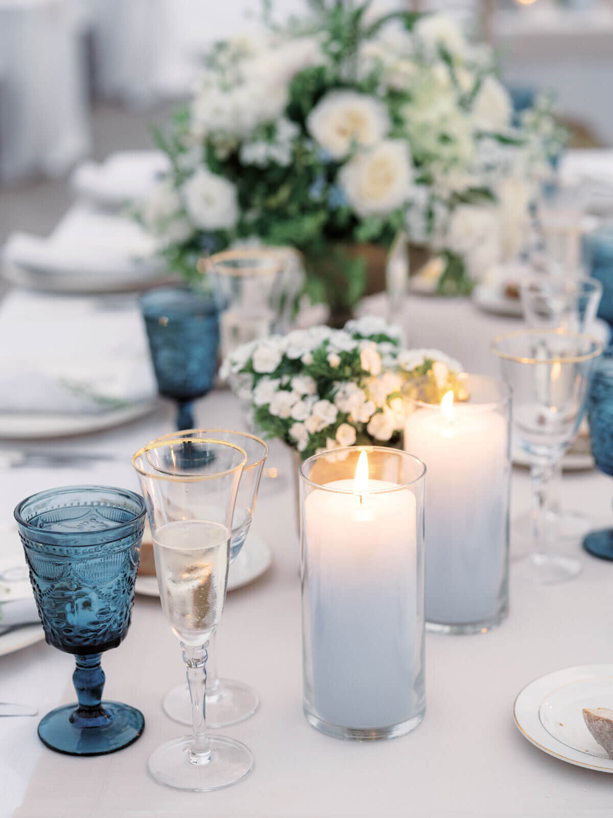 Big white candles at an elegant wedding dining table with flowers, cutleries, and blue wine glasses. Image by Jenny Fu Studio