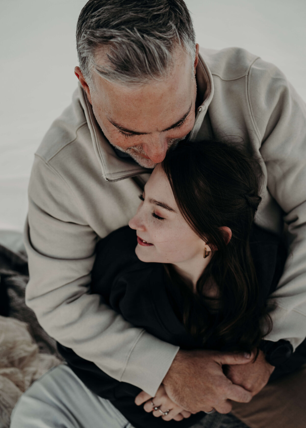 A father kisses, his teenage daughters for head as the audience observes from above. The daughter is wearing a dark sweater, and the father is wearing a cream colored sweater with a snowy backdrop.