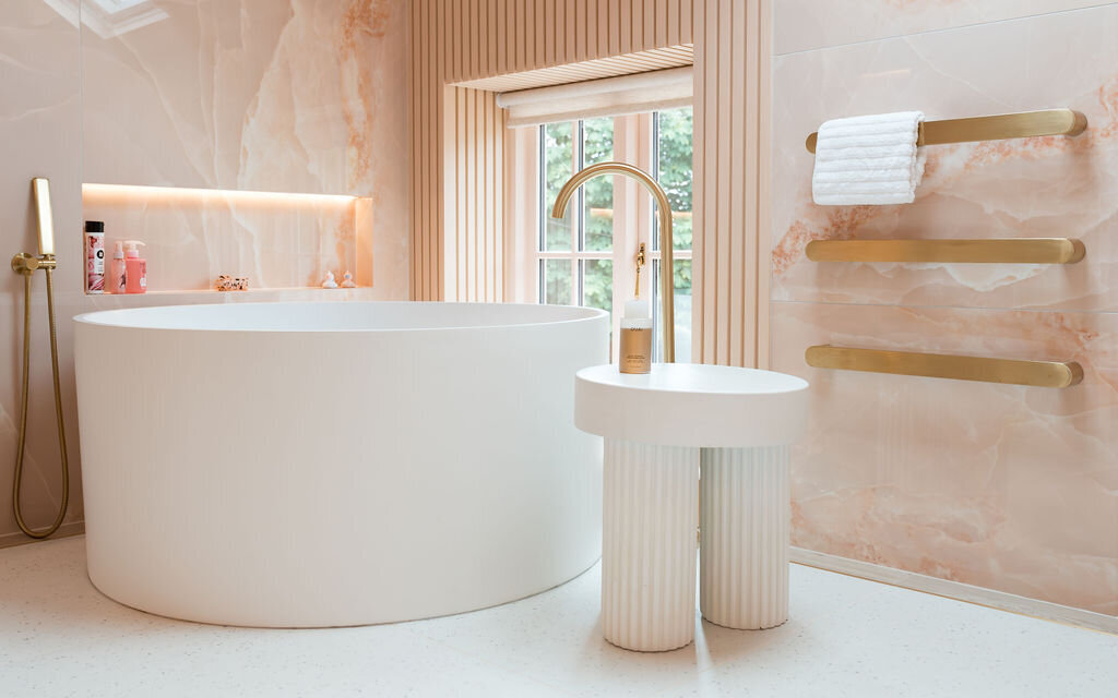 A modern bathroom with a round white bathtub, a sleek gold faucet, and a matching gold showerhead. A small white table stands next to the tub. Three white towels hang on elegant gold bars mounted on a pink marble wall. A window with vertical blinds adds natural light.