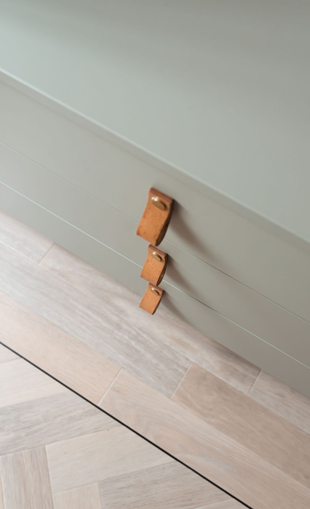 Close-up details of a pastel green dresser in a cozy children's playroom. The dresser sits on light herringbone flooring and has brown leather pulls.