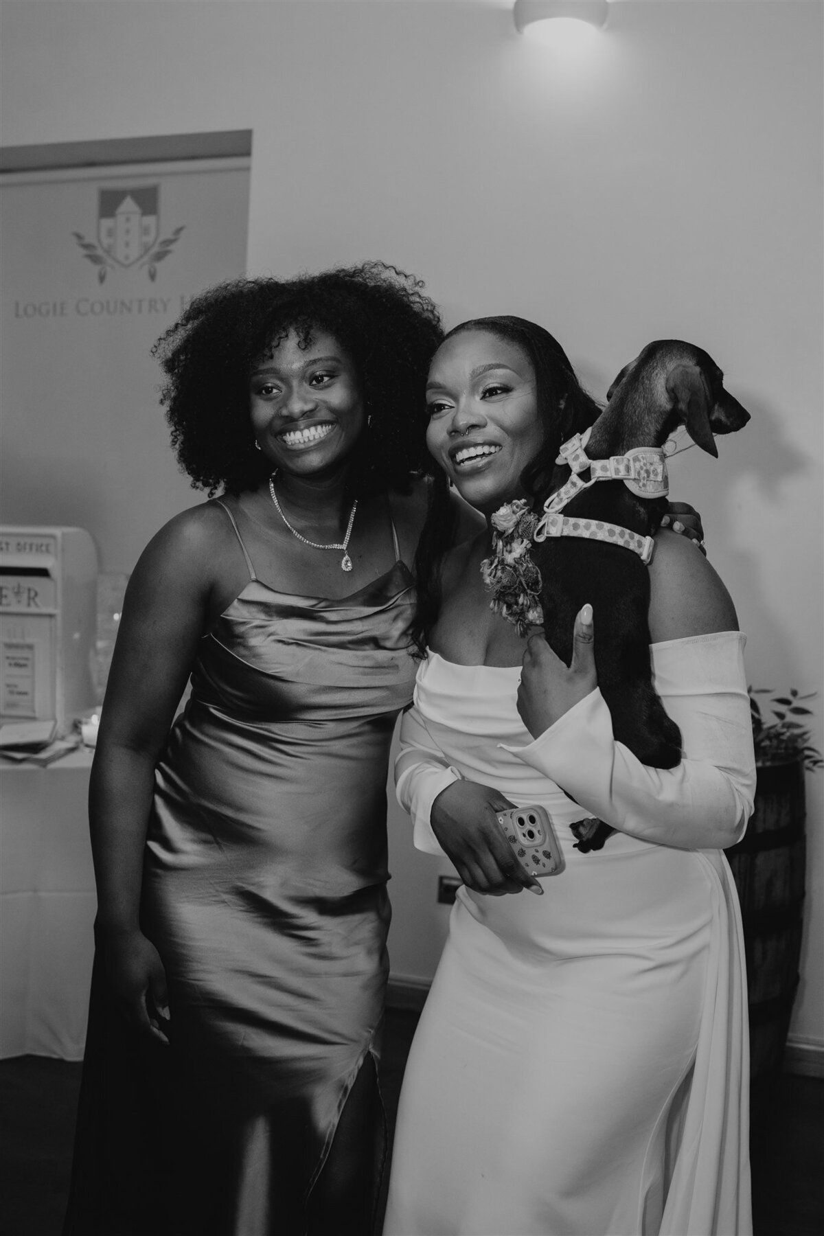 A bride holds a small dog and poses with a guests as another guests takes their photograph.
