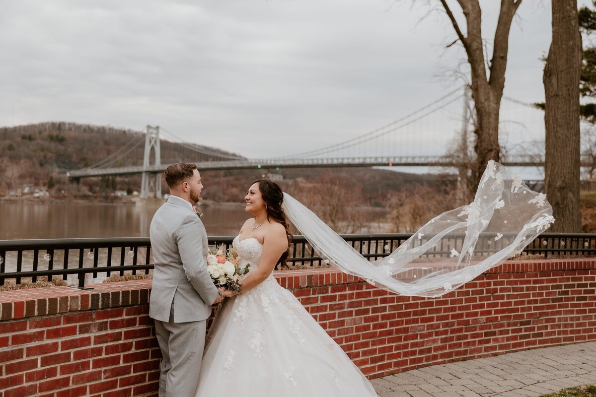 Bride and groom smiling at each other during their wedding portrait at The Grandview in Poughkeepsie, with the Mid-Hudson Bridge and Hudson River in the background. The bride’s veil flows gracefully in the breeze.