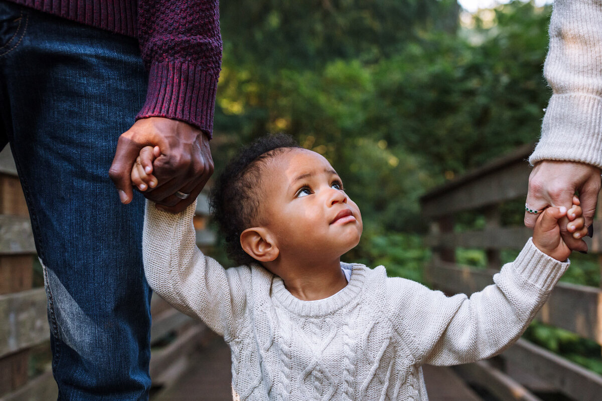 toddler boy standing in a Berkeley park between both parents and holding each of their hands as he looks up as his mom