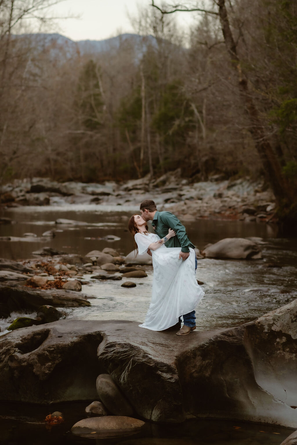 Groom dipping and kissing bride on a large rock by the river at Greenbrier during their eloping to Gatlinburg session, with a mountain backdrop and bare trees suggesting late fall or early winter.
