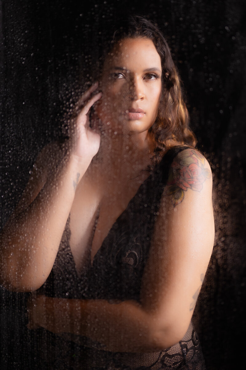 a black woman in a black lacy bodysuit stands behind the wet and steamy shower door where there are water droplets.