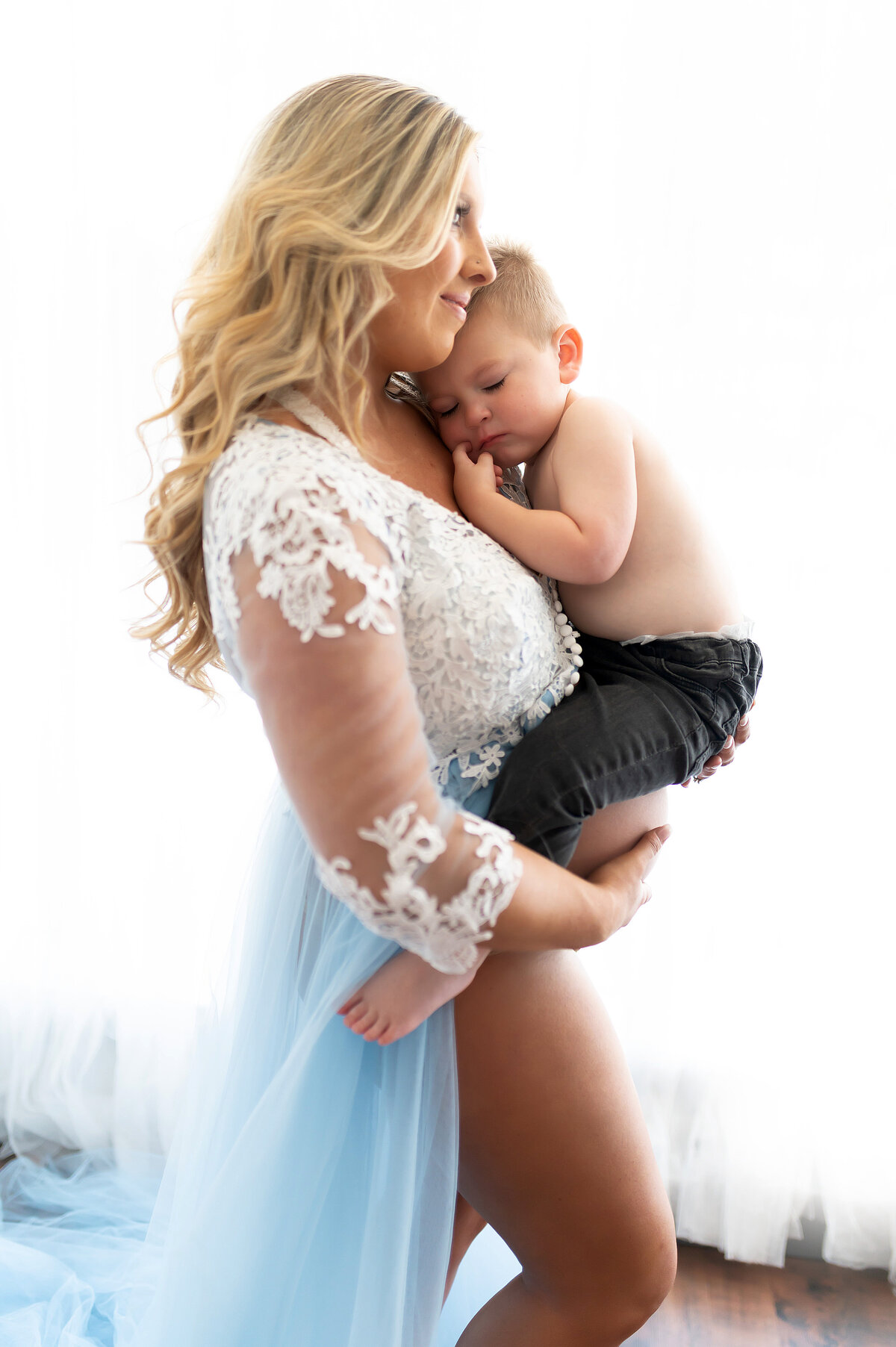 Pregnant mother in a blue tulle gown with lace bodice holding her toddler son, captured in soft natural light during a studio maternity session in Waukesha, Wisconsin.