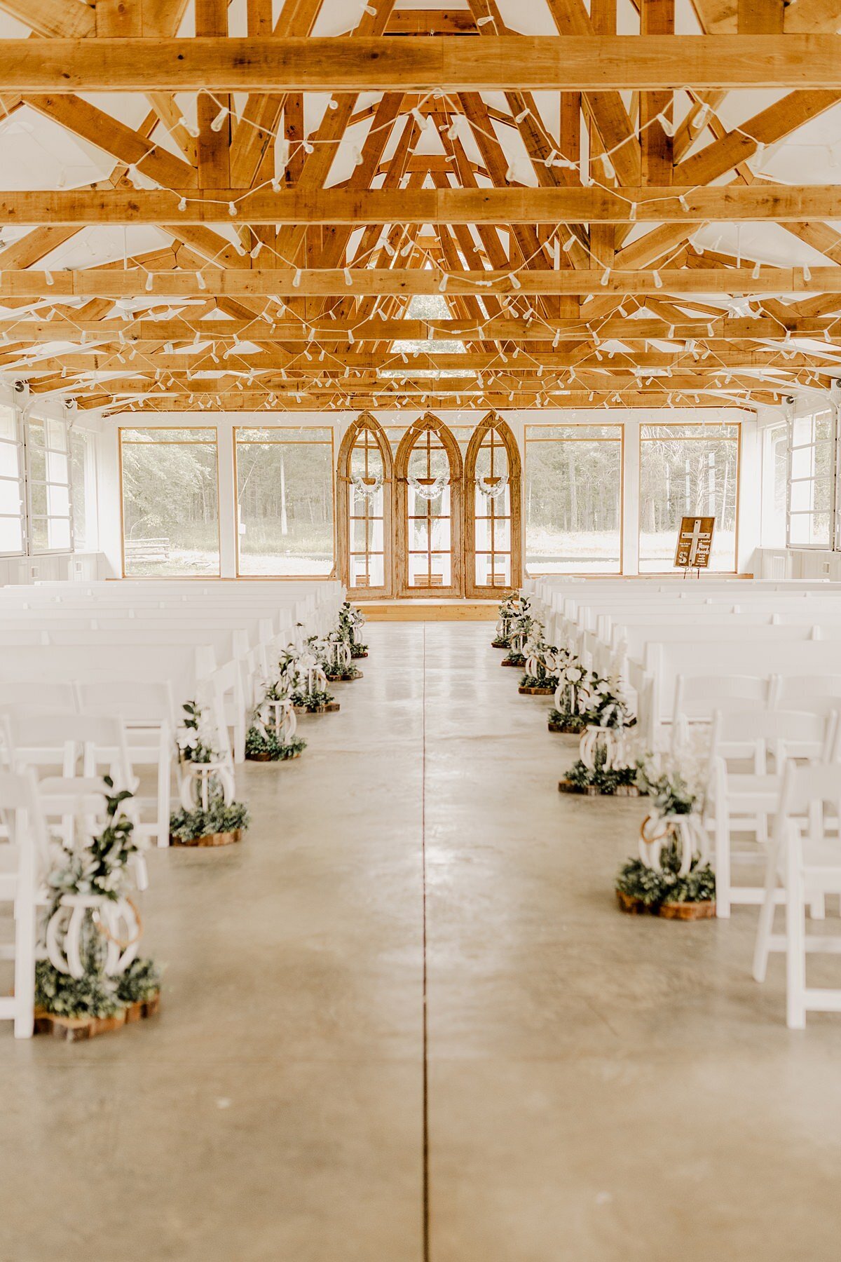 A greenhouse with exposed wood beams wrapped with white twinkle lights is set with white garden chairs for a wedding. The aisle is lined with white lanterns decorated with eucalyptus. A three piece gothic window is set at the end of the aisle at Grace Valley Farm.