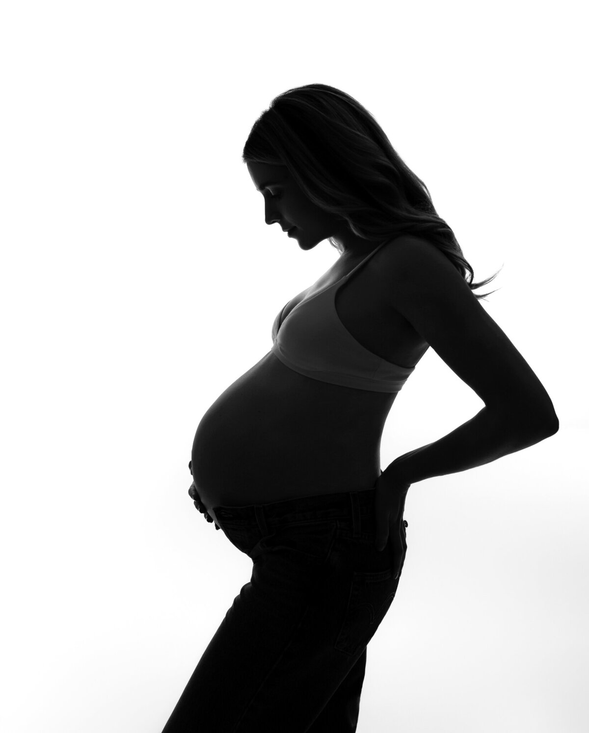 Modern maternity silhouette photo of a mom-to-be in jeans and a crop top, captured with high-contrast studio backlighting.