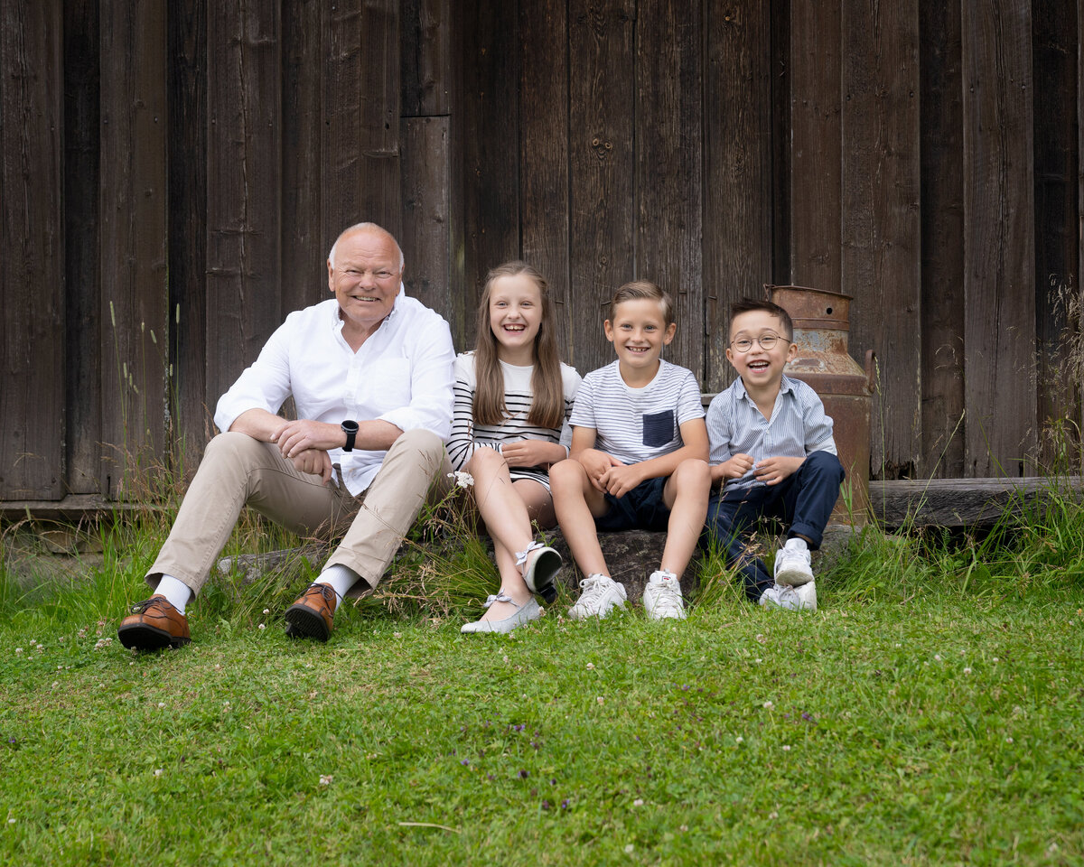 Bestefar og tre barnebarn sitter sammen på gress foran en rustikk trevegg under familiefotografering nær Oslo.
