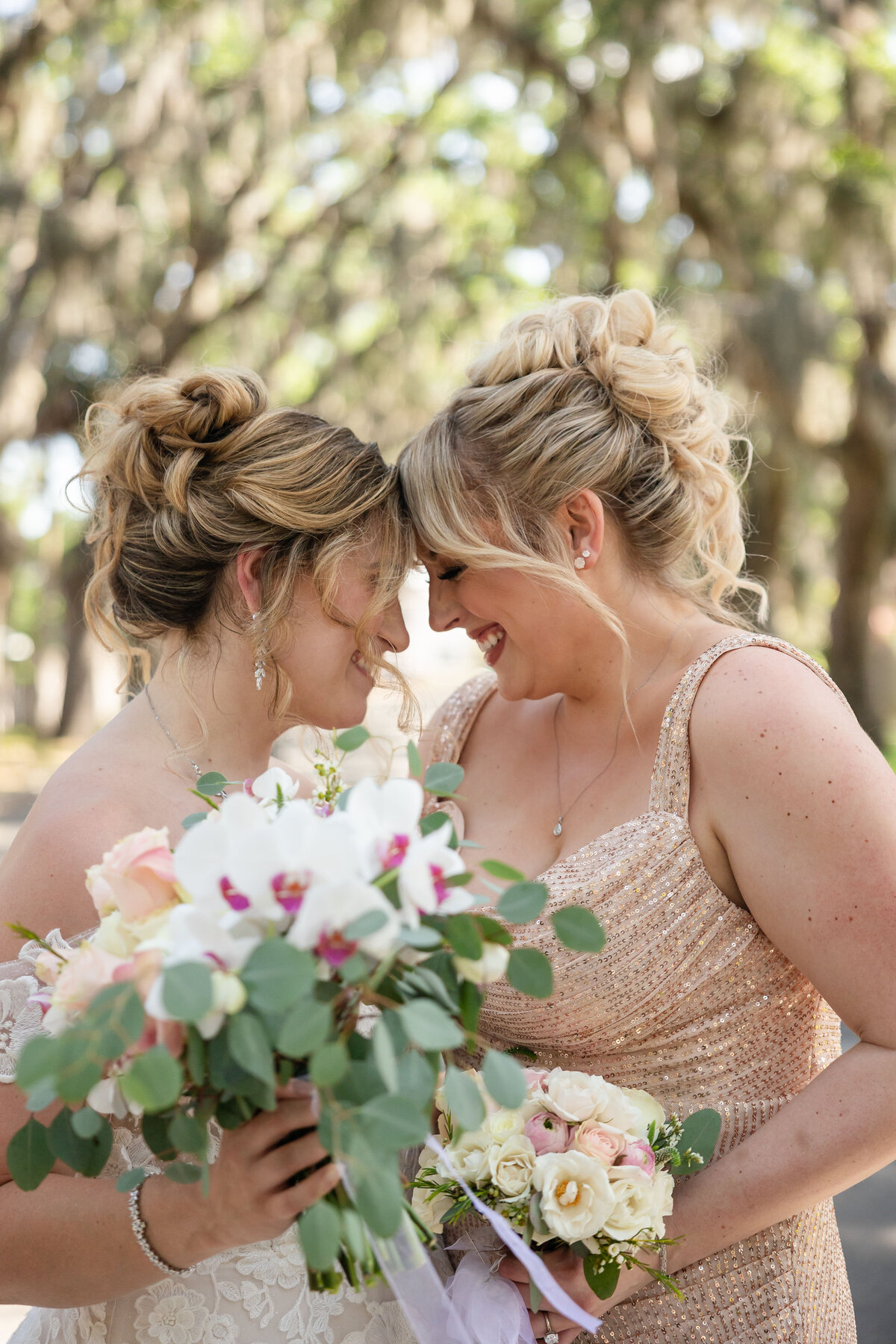 The bride and her maid of honor lean in with their foreheads touching, laughing together during a sweet moment at the Fountain of Youth in St. Augustine, Florida. Surrounded by soft light and moss-draped trees, the bride holds a bouquet of white orchids and pink roses. The maid of honor wears a sparkly blush gown, complementing the joyful, elegant atmosphere. Captured by a Jacksonville wedding photographer who focuses on heartfelt, candid wedding moments.