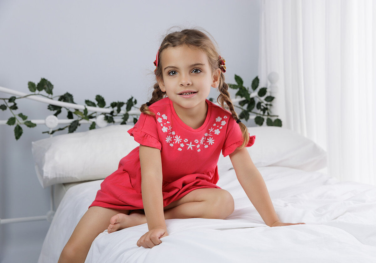Image of a girl sitting on a white bed with holly on the headboard Christmas backdrop in the studio. Taken by Norwich portrait photographer Claire Howes. Christmas Mini Sessions Norwich.