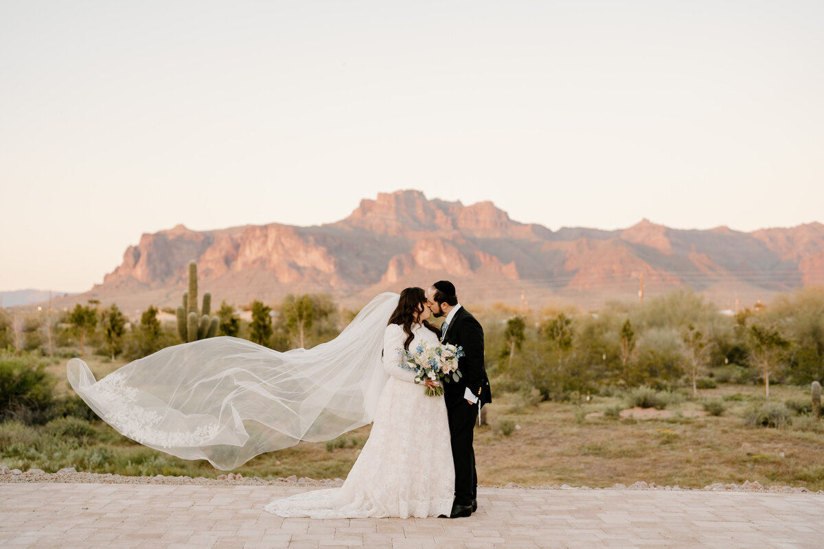 bride and groom kissing in front of superstition mountains with her veil blowing in wind