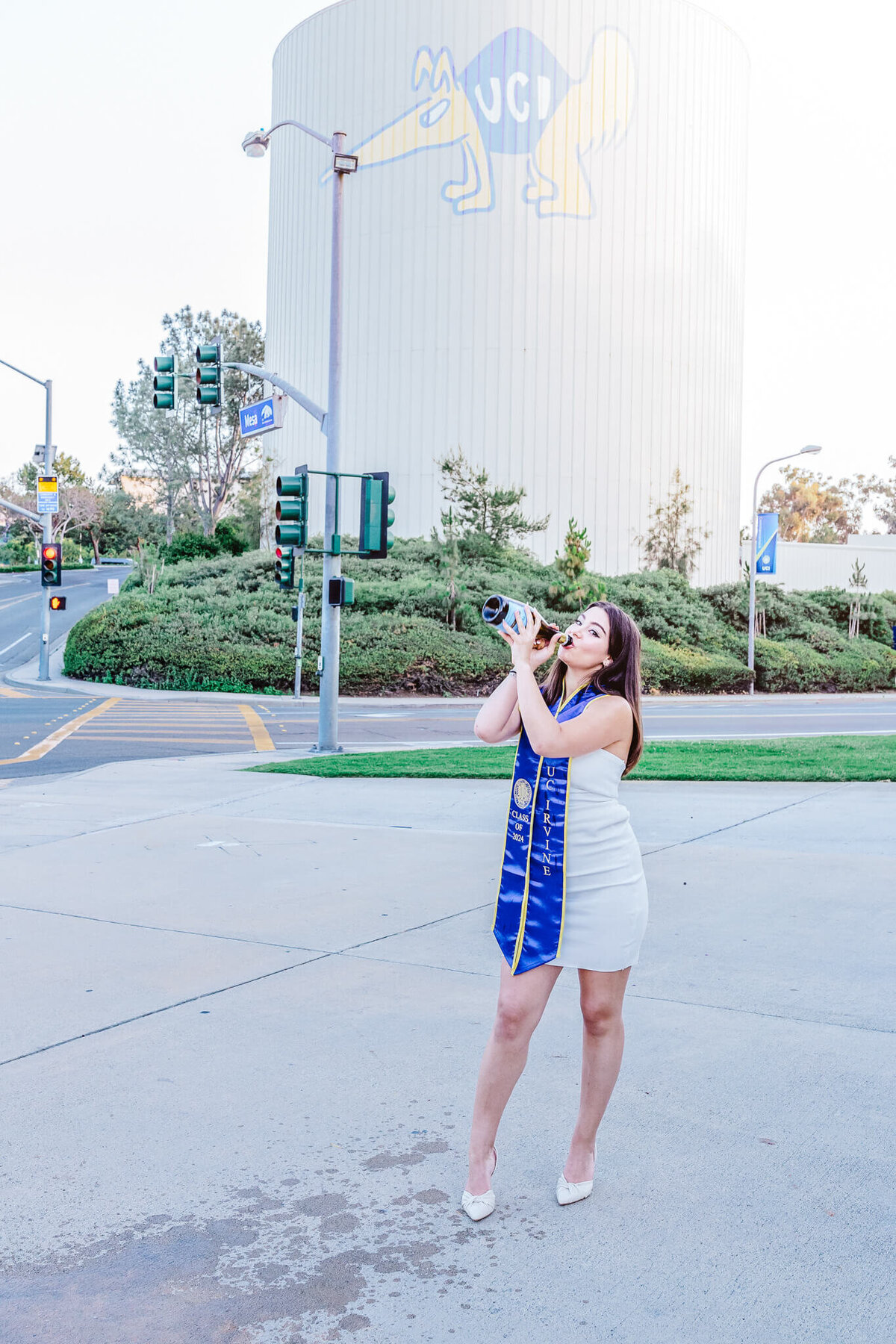 A woman in a white dress and blue graduation stole drinks from a champagne bottle in front of a water tank with a university logo. She stands on an empty street with traffic lights and green foliage in the background.