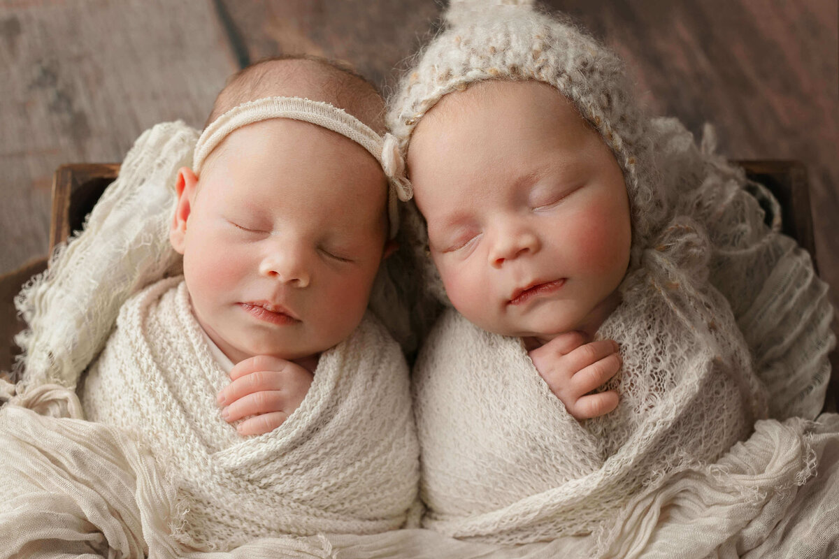 newborn twins in cream colored wraps leaning their heads together in a box at a newborn photo shoot