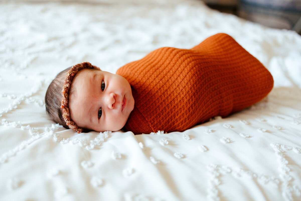 Photo of a newborn baby girl looking towards the camera, she has an orange blanket and is on a white mattress