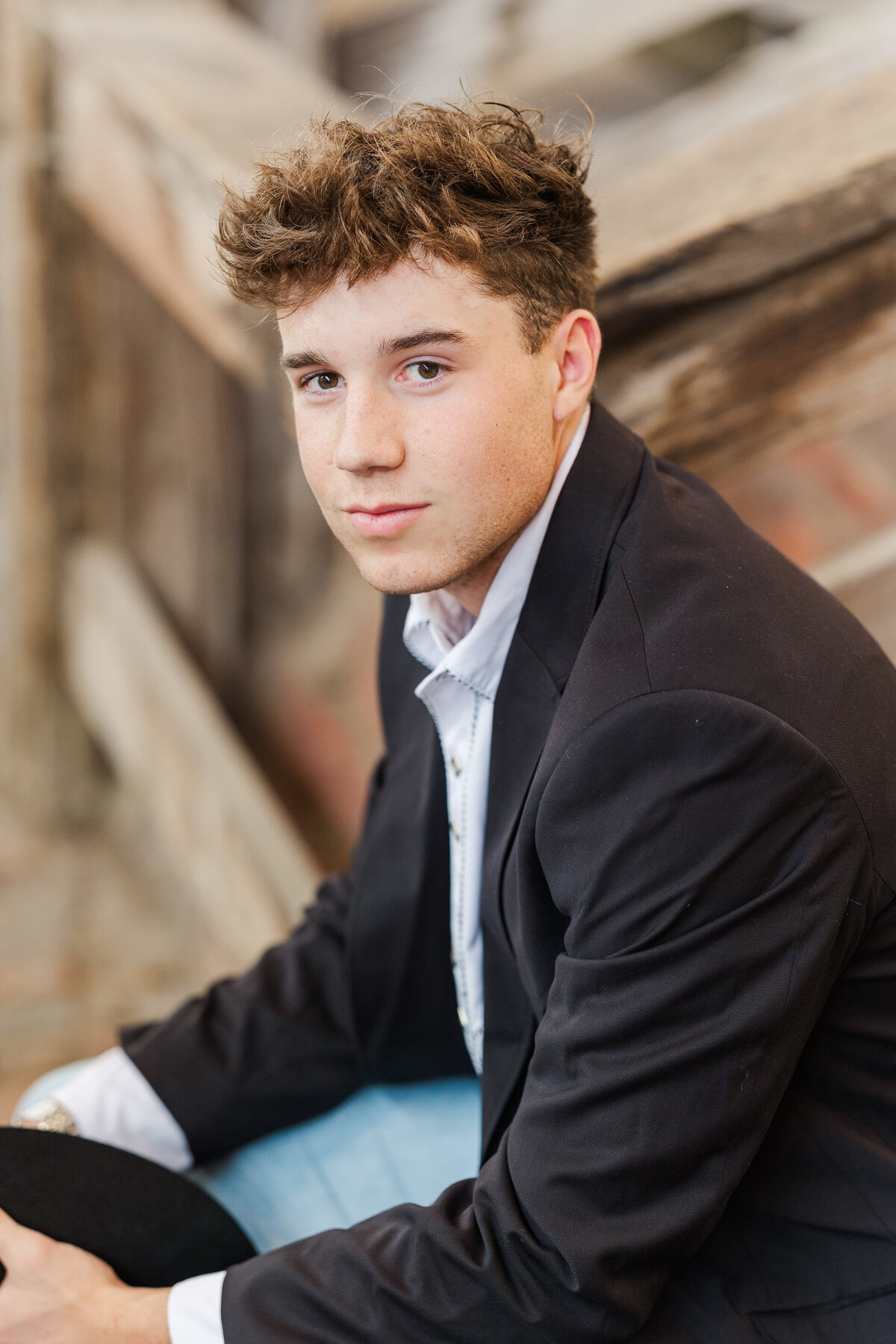 Senior boy sitting on rustic wooden steps in the Fort Worth Stockyards, wearing a black blazer over a white button-up shirt. He looks confidently toward the camera with a slight smile, capturing a rugged and polished Western style.