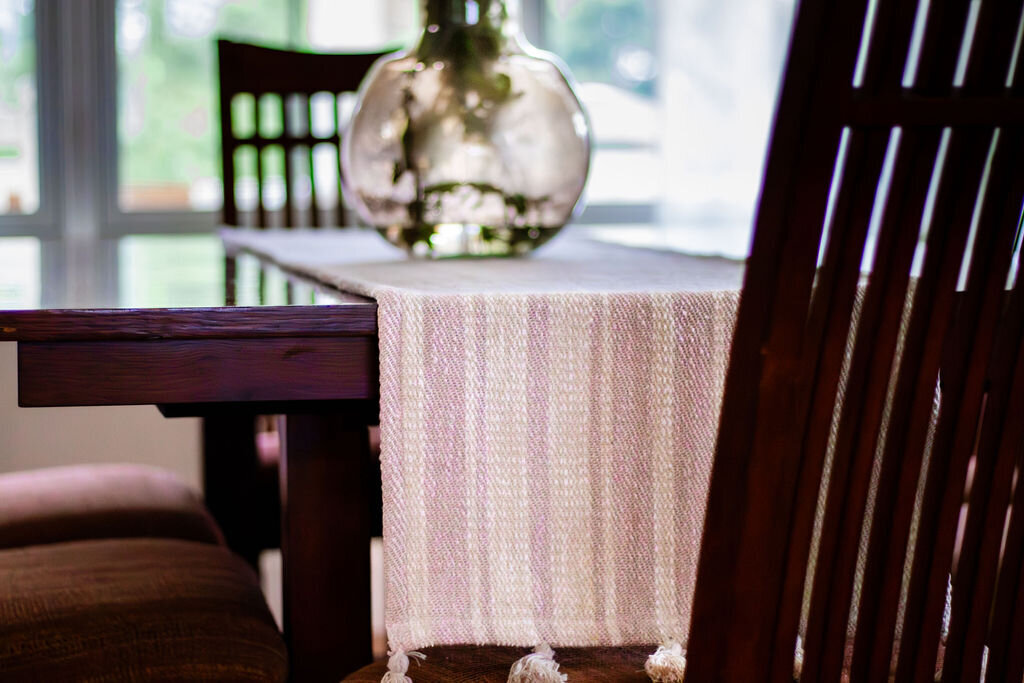 A close-up of a dark wooden dining table with a light-colored, vertically striped table runner featuring tassels at the end. A clear glass vase, slightly out of focus, sits on the runner. The backs of two dark wooden chairs with vertical slats are partially visible, one in the foreground and another behind the table. Soft light comes from a window in the background.