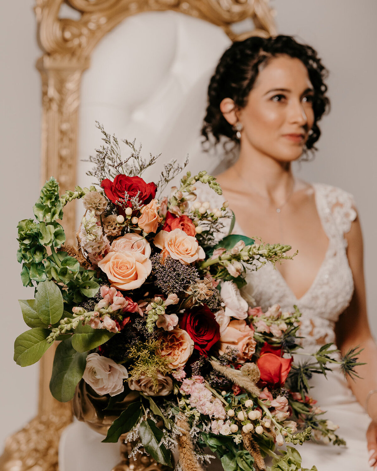 The bride, seated on an ornate white and gold chair, holds a large, colorful bouquet of red, peach, and pink flowers.