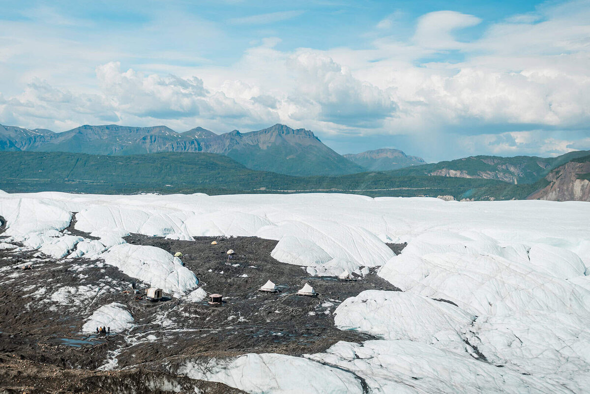 The view of on-glacier glamping tents as seen approaching by private helicopter during a glacier elopement wedding package .  The luxury tents look like three tiny white dots on the dark colored rocky glacier moraine that sits between the mounts of white glacier ice. 