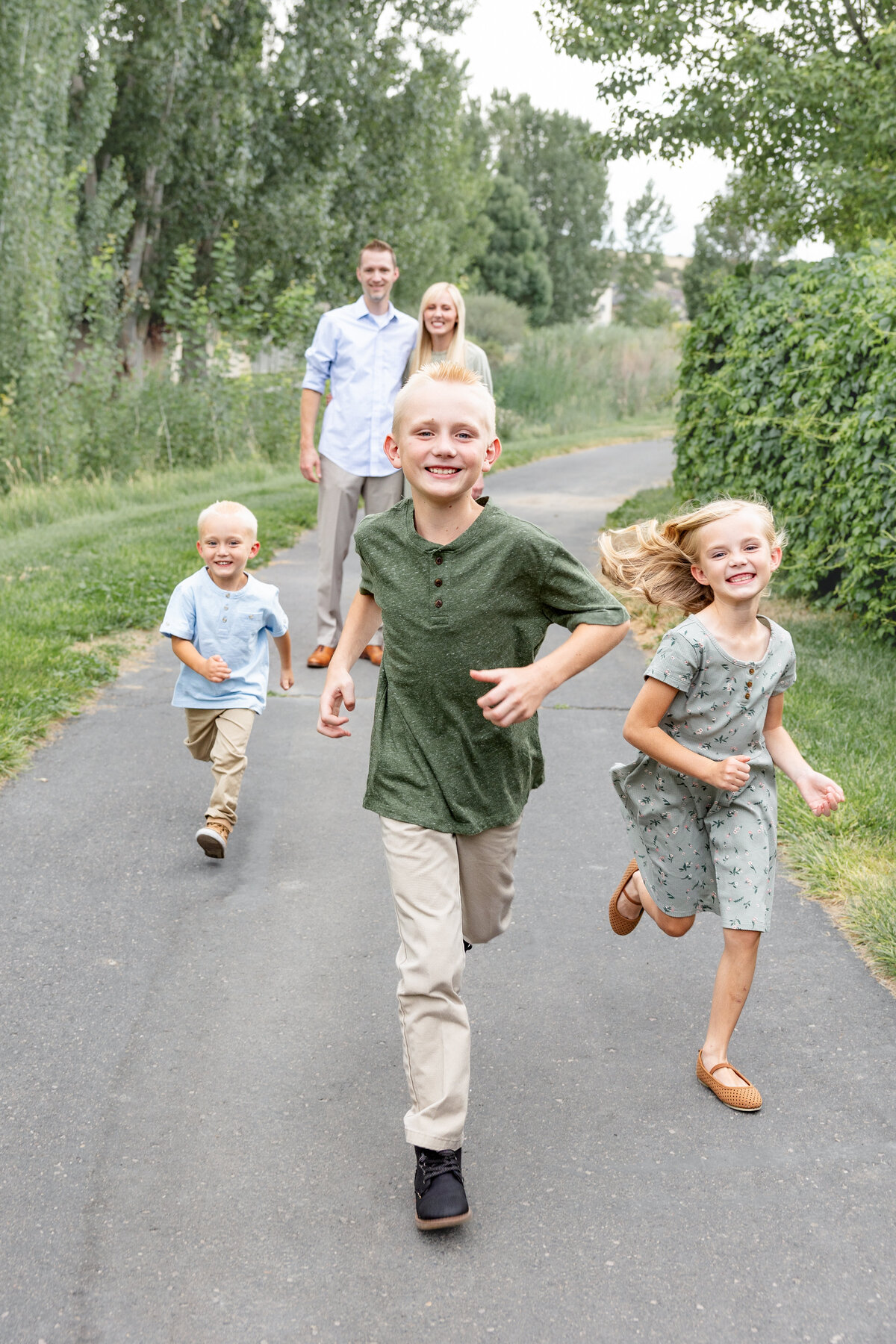 Three smiling children running toward the camera on a paved path, with their parents in the background.