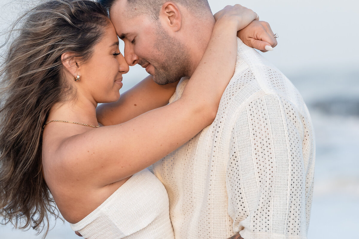 Man and woman both wearing white holding each other foreheads touching with their eyes closed as the wind on the beach blows her hair.