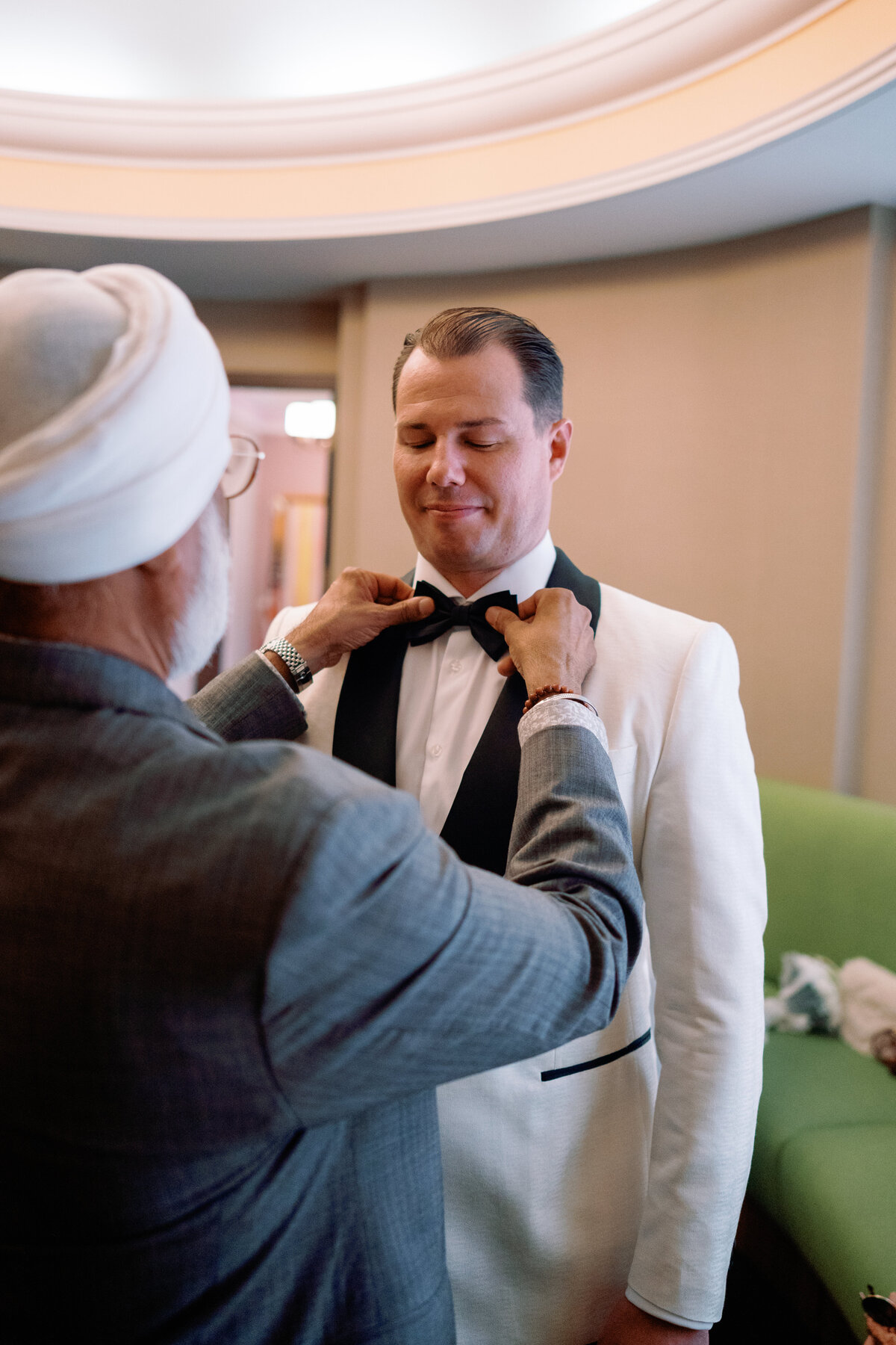 Groom smiling as his father helps adjust his bow tie inside New York City Hall before the ceremony, captured during Japna and Chris’s intimate elopement by NYC wedding photographer Perry Hancock.