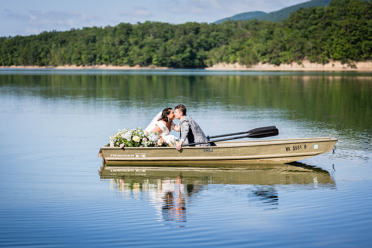 A couple go for a kiss in a rowboat in the waters of Carvin’s Cove with the rolling hills of Southwest Virginia in the background. The boat is adorned with pastel flowers and a sign that reads “just married” on the back of the boat. The reflection of the couple kissing can be seen in the water.