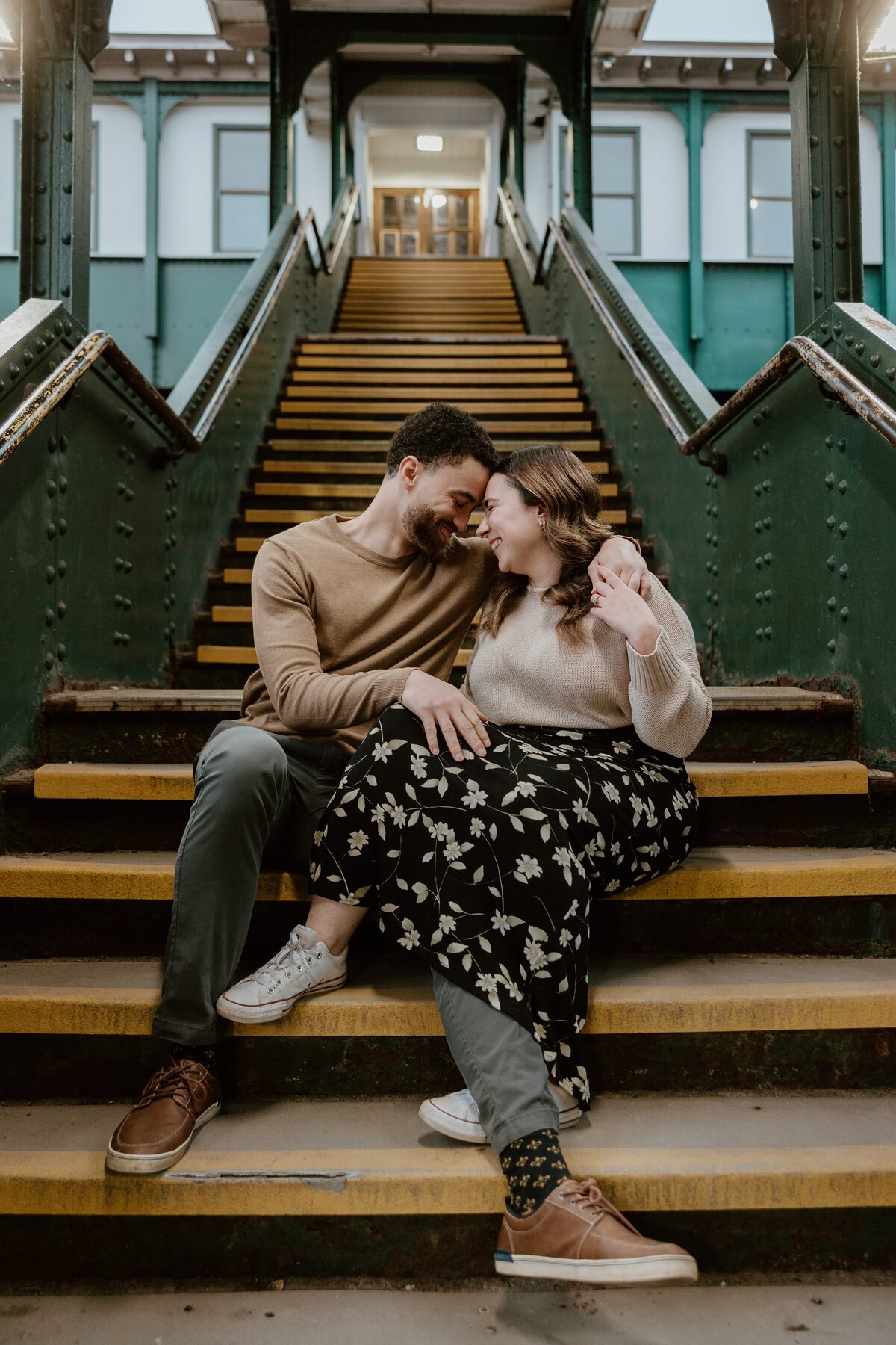 A couple sitting close together on the yellow-striped steps of the Poughkeepsie Train Station, sharing a warm, candid moment. They are smiling with their heads touching, conveying a sense of comfort and affection. The setting features the station’s rustic green metal railings and industrial architecture, enhancing the cozy and intimate atmosphere of the photo.