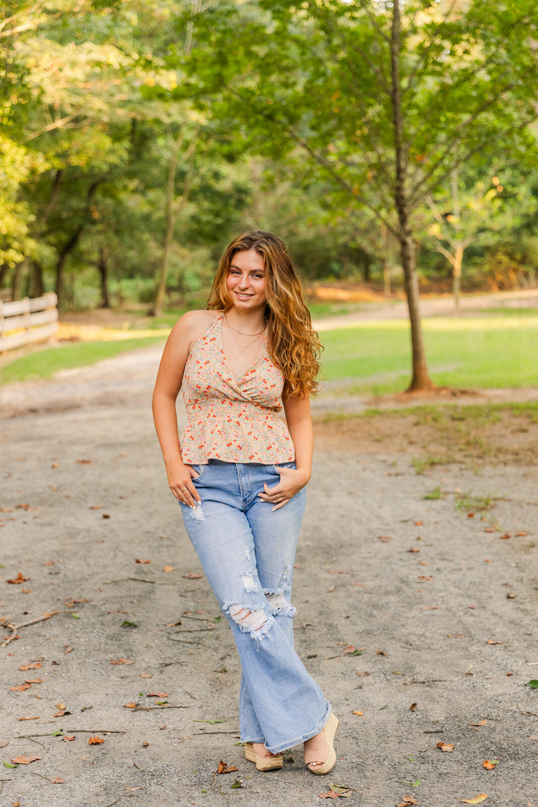 wide shot of senior portrait standing on a park path ands in jeans pockets  and ankles crossed