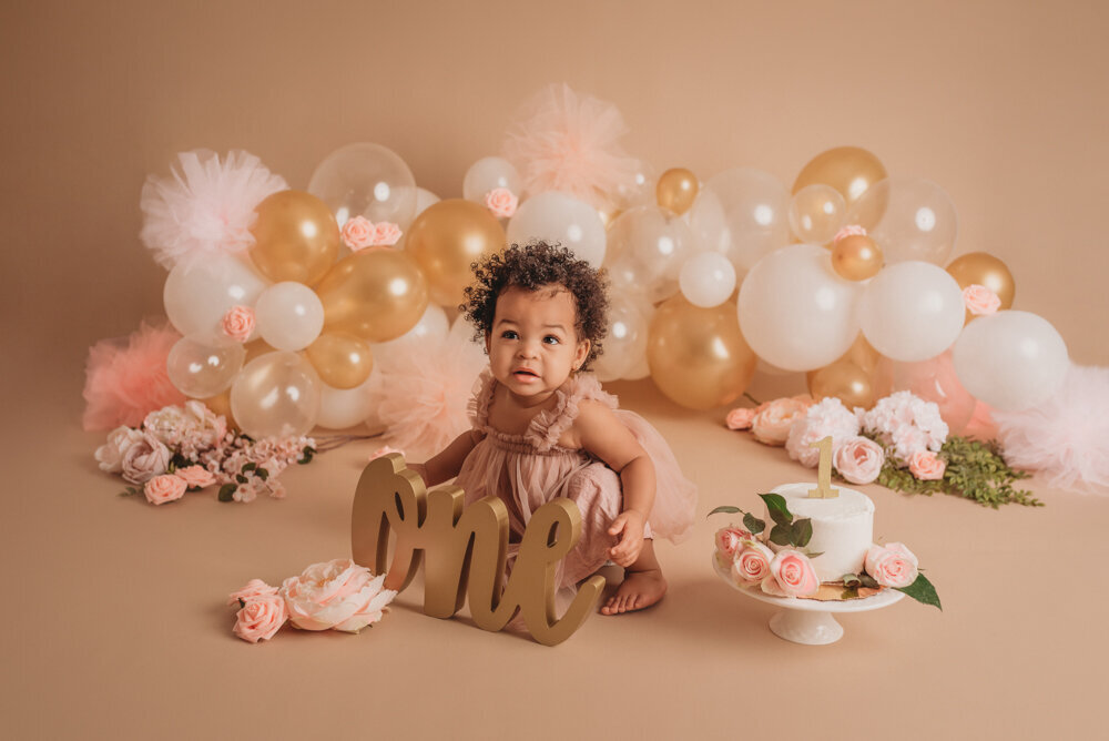 Cake smash session with Atlanta, Georgia cake smash photographer of one year old baby girl in pink tulle dress on tan background with neutral colored balloon garland eating her white iced birthday cake with pink roses on it