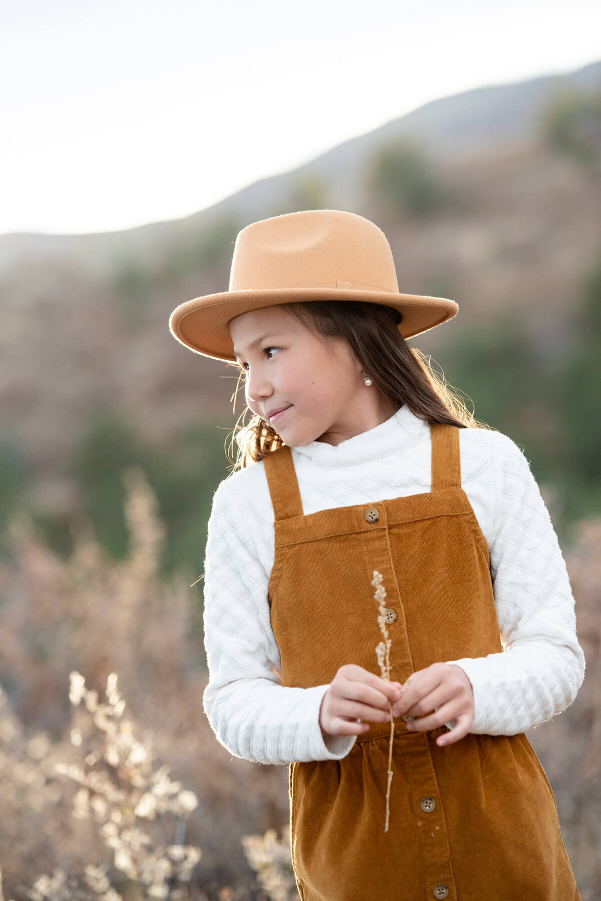 A young girl in an orange dress and brown hat holds a piece of grass while exploring a mountain trail