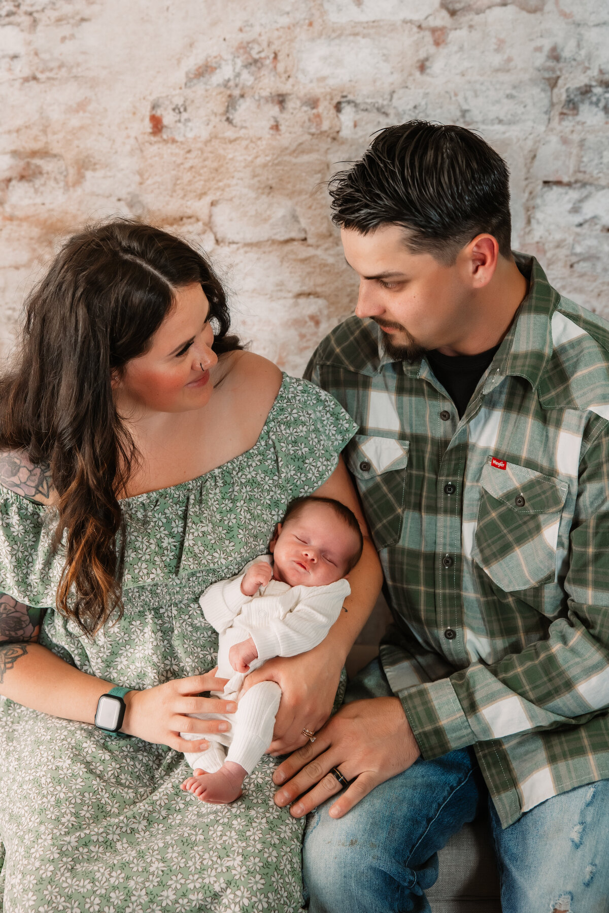 Happy mom and dad sit on a bench in a studio cradling their newborn baby between them