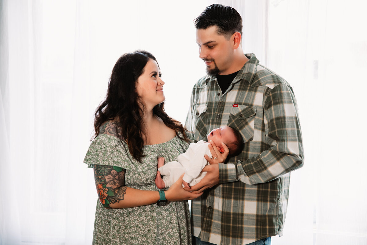 Happy new mom and dad stand in green outfits in a studio holding their sleeping newborn baby together