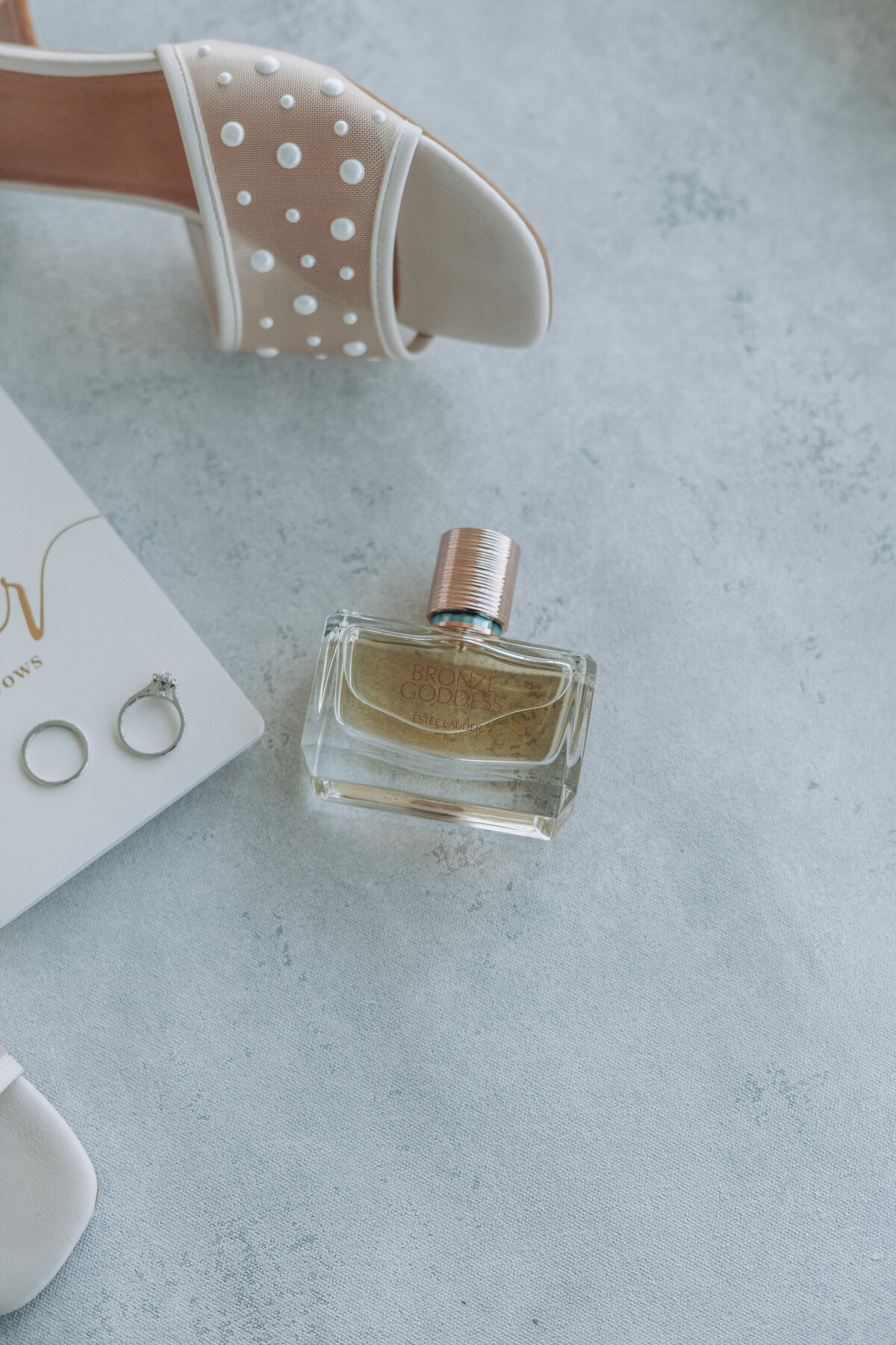 Close-up of delicate wedding details including a bride's perfume bottle, sparkling wedding rings, and a bridal shoe, beautifully arranged at the Lesner Inn in Virginia Beach, Virginia, captured by Quinn Photography LLC.