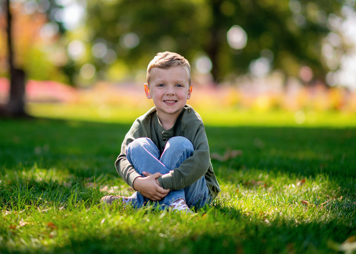 A boy in a green shirt sits in the grass at a park with trees