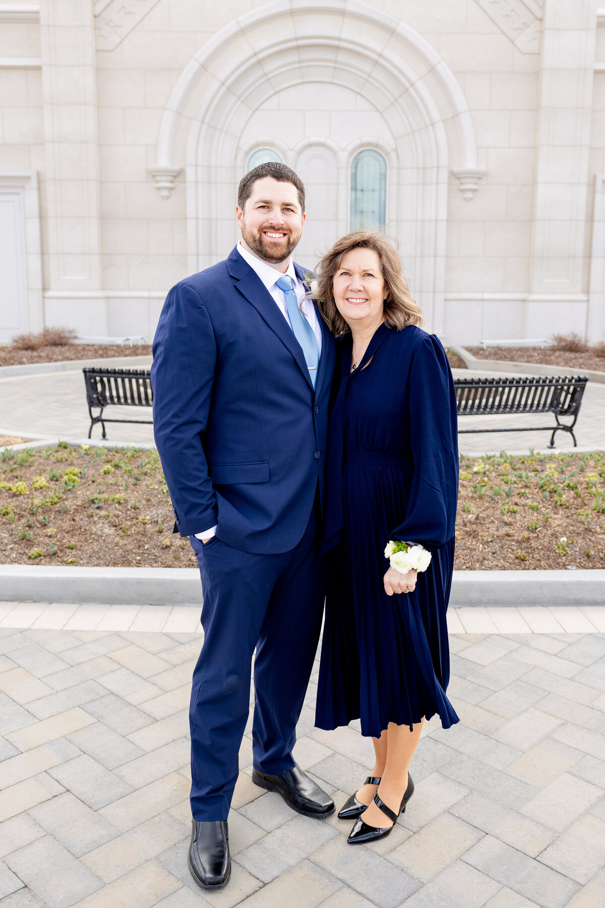 Groom stands closely with his mother, both smiling warmly and wearing matching navy blue attire, in front of the LDS temple.