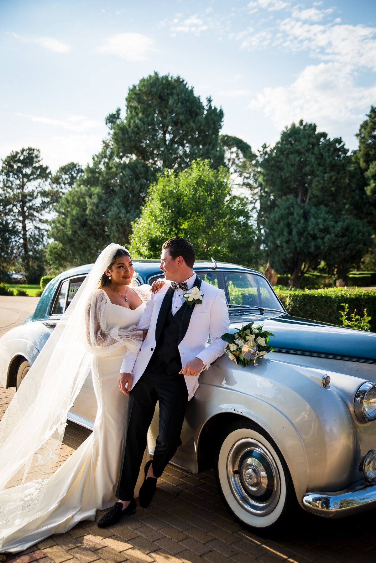 A groom leans on a silver vintage getaway car as his bride stands close, holding his arm.