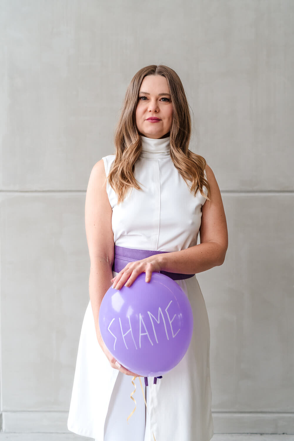 creative headshot for entrepreneur wearing white outfit and holding purple balloon with the word shame on it front of her belly