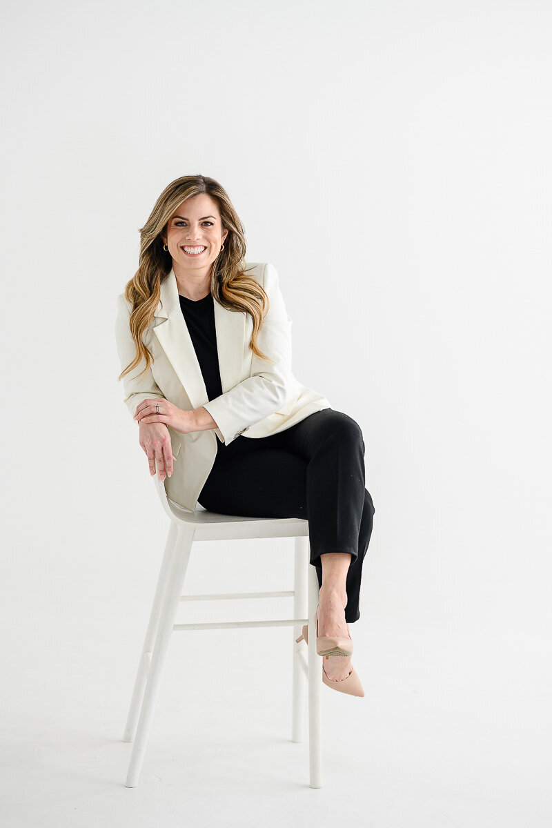 A branding photographer in Denver takes a picture of a real estate agent in a white suit sitting on a white stool for her branding photos and brand photography photo shoot.