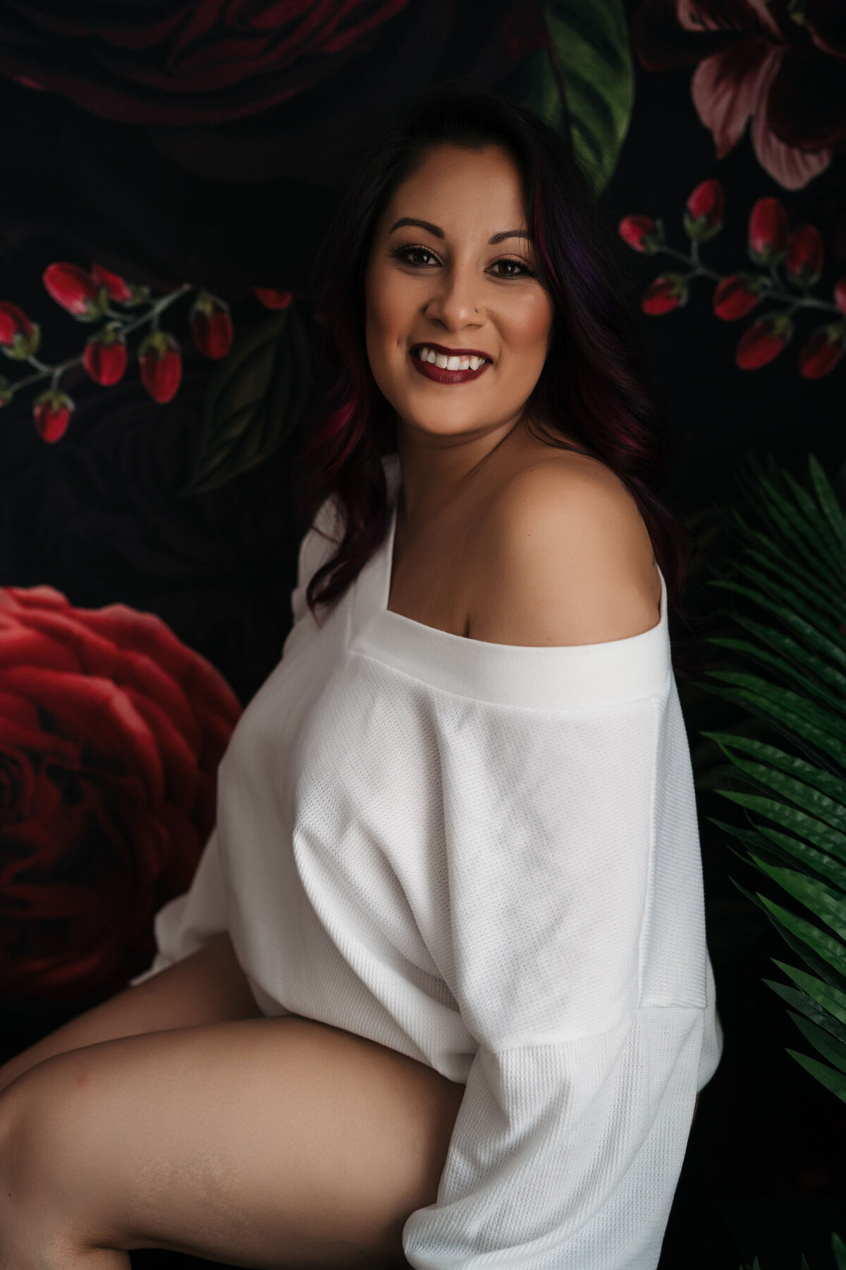 A woman smiles while sitting in a white oversized shirt in front of a large floral tapestry