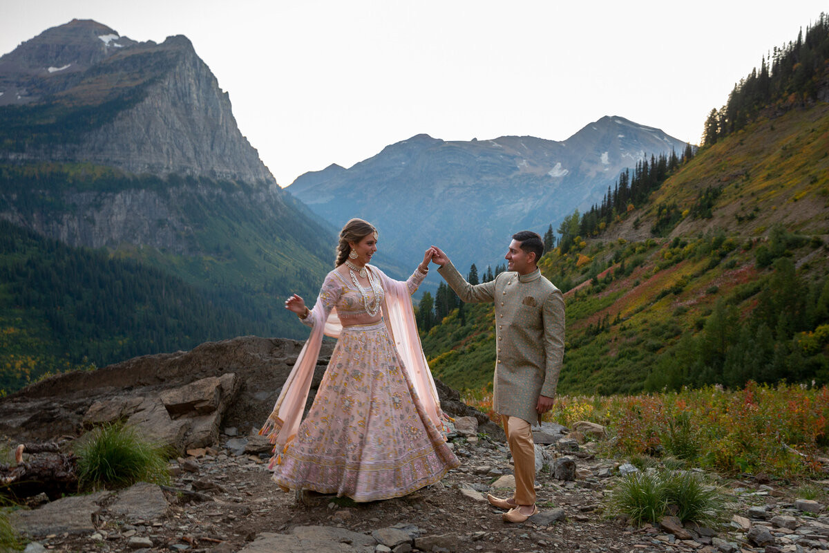 A bride and groom stand on a rocky path in Glacier National Park, dressed in Indian wedding attire and as the groom holds the brides hand.