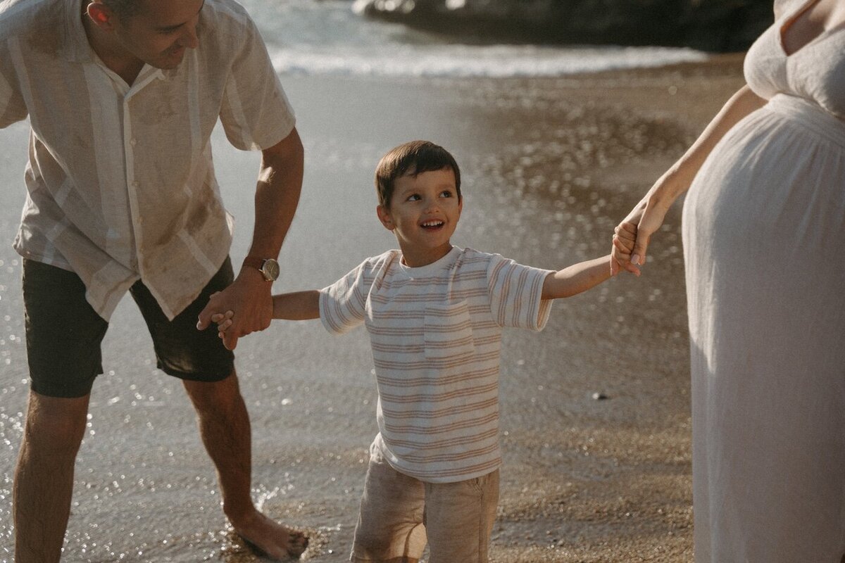 Le petit garçon sourit en tenant la main de ses parents, pieds dans l'eau. Shooting famille par Kamescop, photographe en Loire-Atlantique.