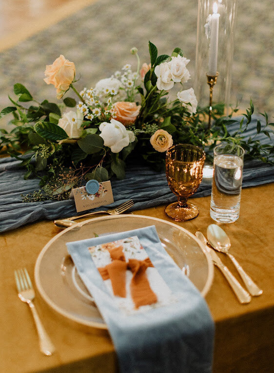A place setting featuring mustard and dusty blue velvet linens with a peach floral centrepiece and gold cutlery is set for a wedding at Chateau Laurier in Ottawa Ontario